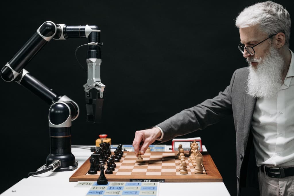 A man with a white beard playing chess against a black industrial robotic arm on a wooden board.